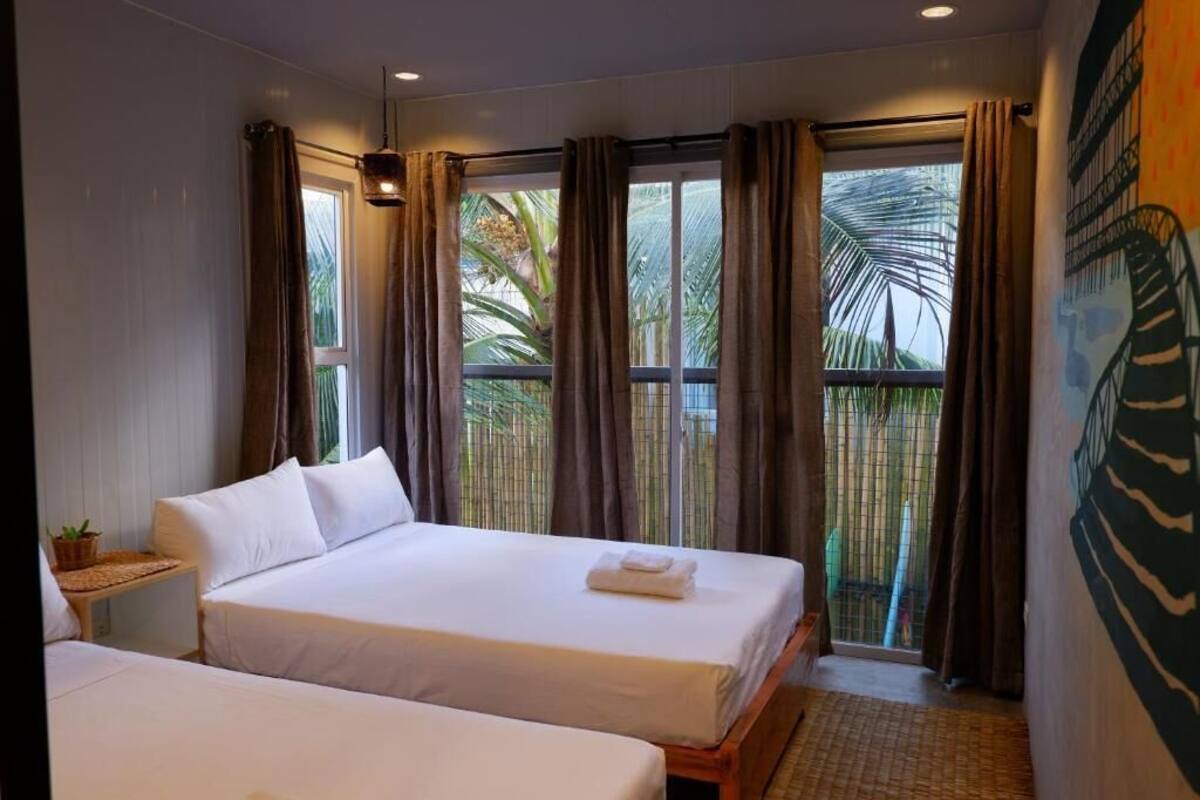 A hotel room with two beds, a small table with a plant, and a view of palm trees through the window.