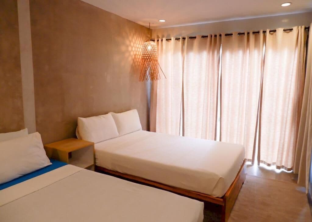 A hotel room with two beds, a wooden nightstand, a wall-mounted lamp, and a large window with curtains.