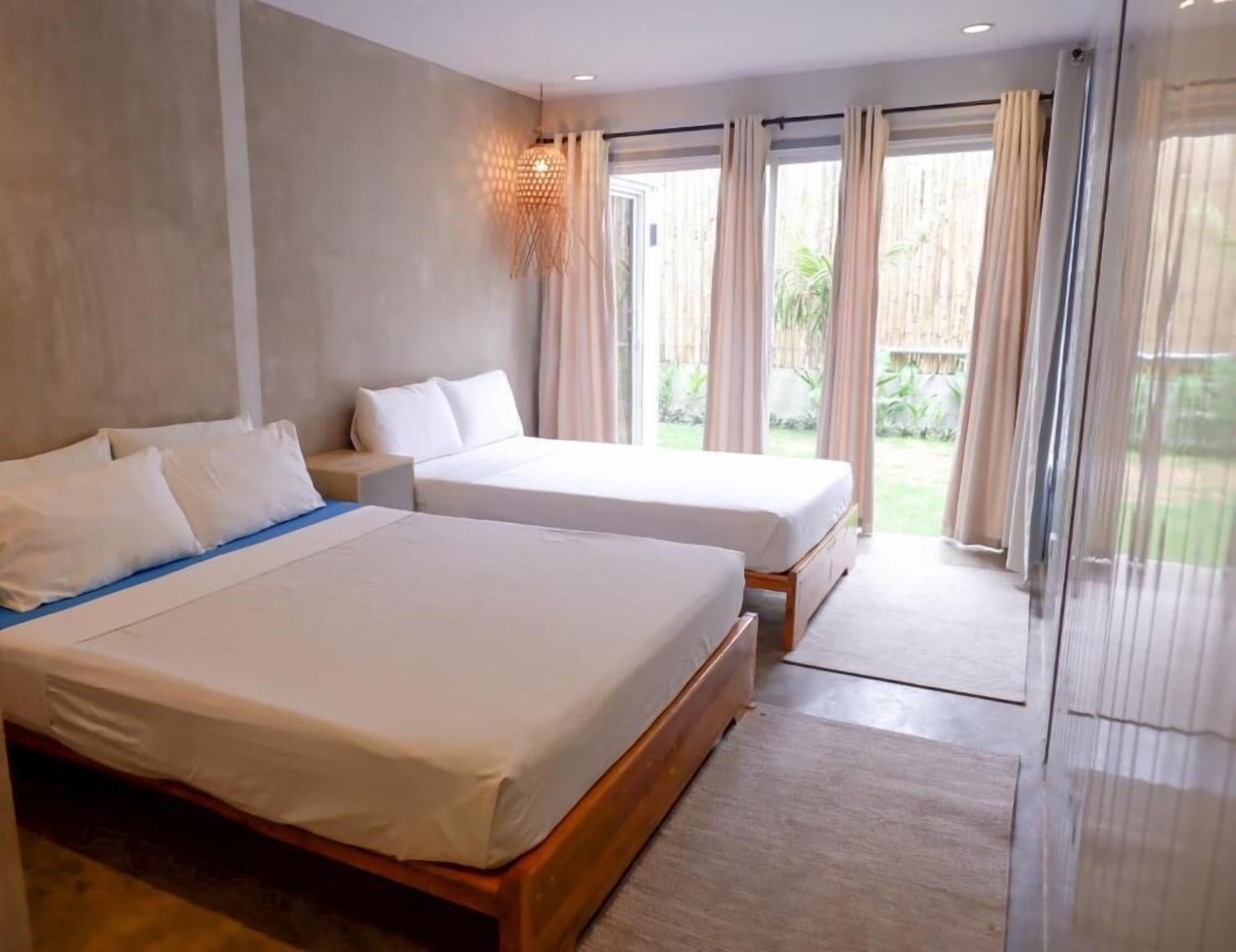 Habitación de hotel con dos camas, una mesita de noche, una lámpara y vistas al exterior.