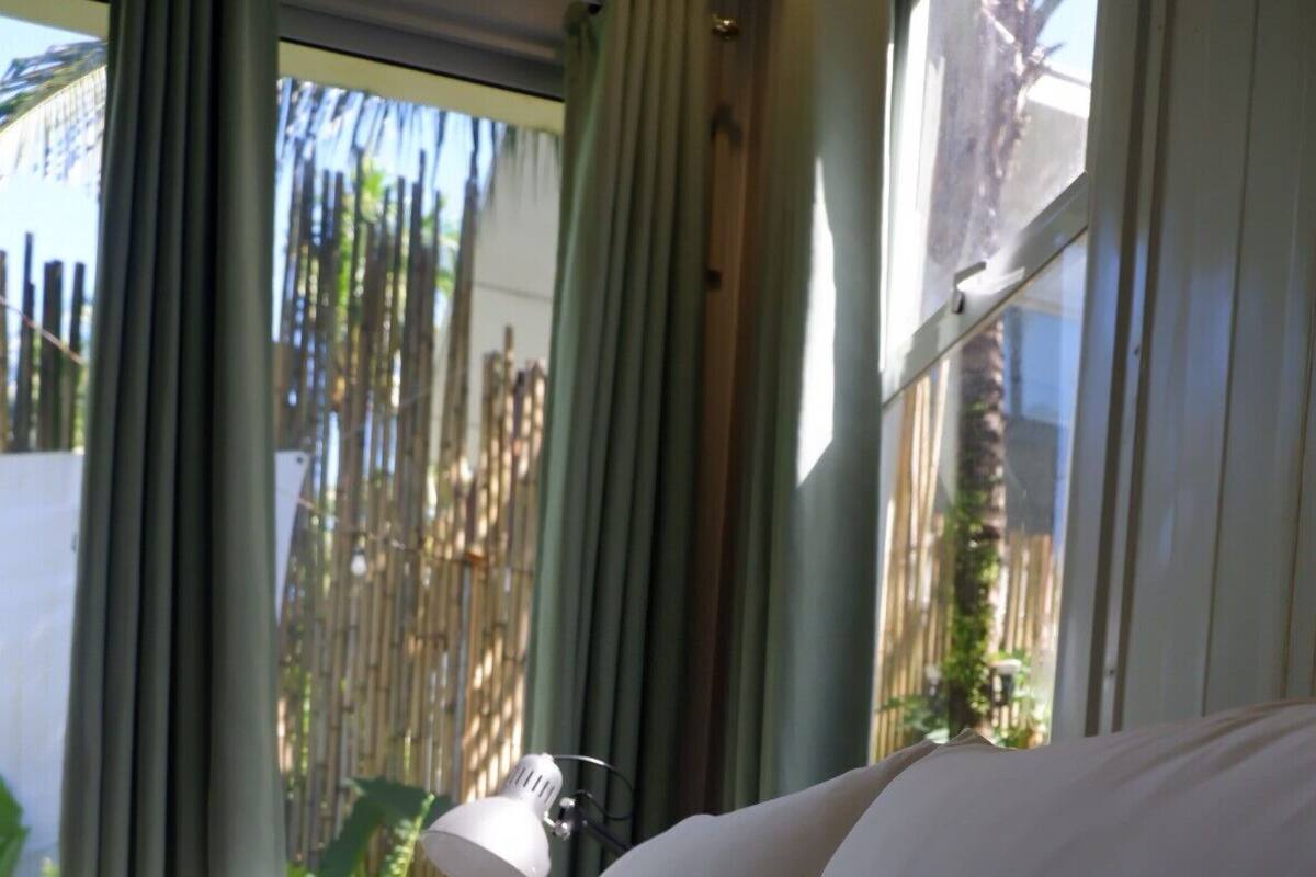 A bed with white linens, a bedside lamp, and a view of a bamboo fence through a window.