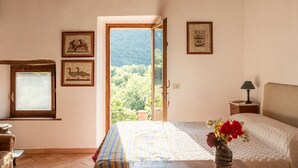 Un dormitori amb un llit, una tauleta de nit, una làmpada, un vas amb flors i una finestra que ofereix vistes a la vegetació.