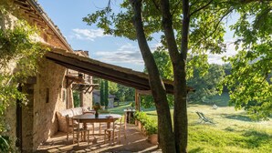 Edifici de pedra rústic amb una pèrgola de fusta, una taula rodona amb caires i un jardí enjardinat amb unes quantes cadires de relax.
