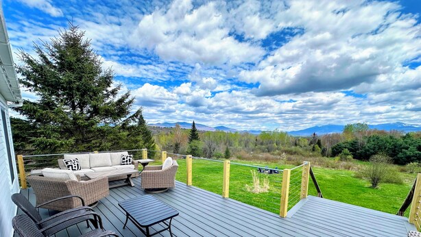 A deck with outdoor furniture, a view of a grassy field, and mountains in the distance. - 64J Stunning sunsets, just 20 mins to Bretton Woods. Hot Tub, Foosball, AC, dog friendly! (Whitefield)