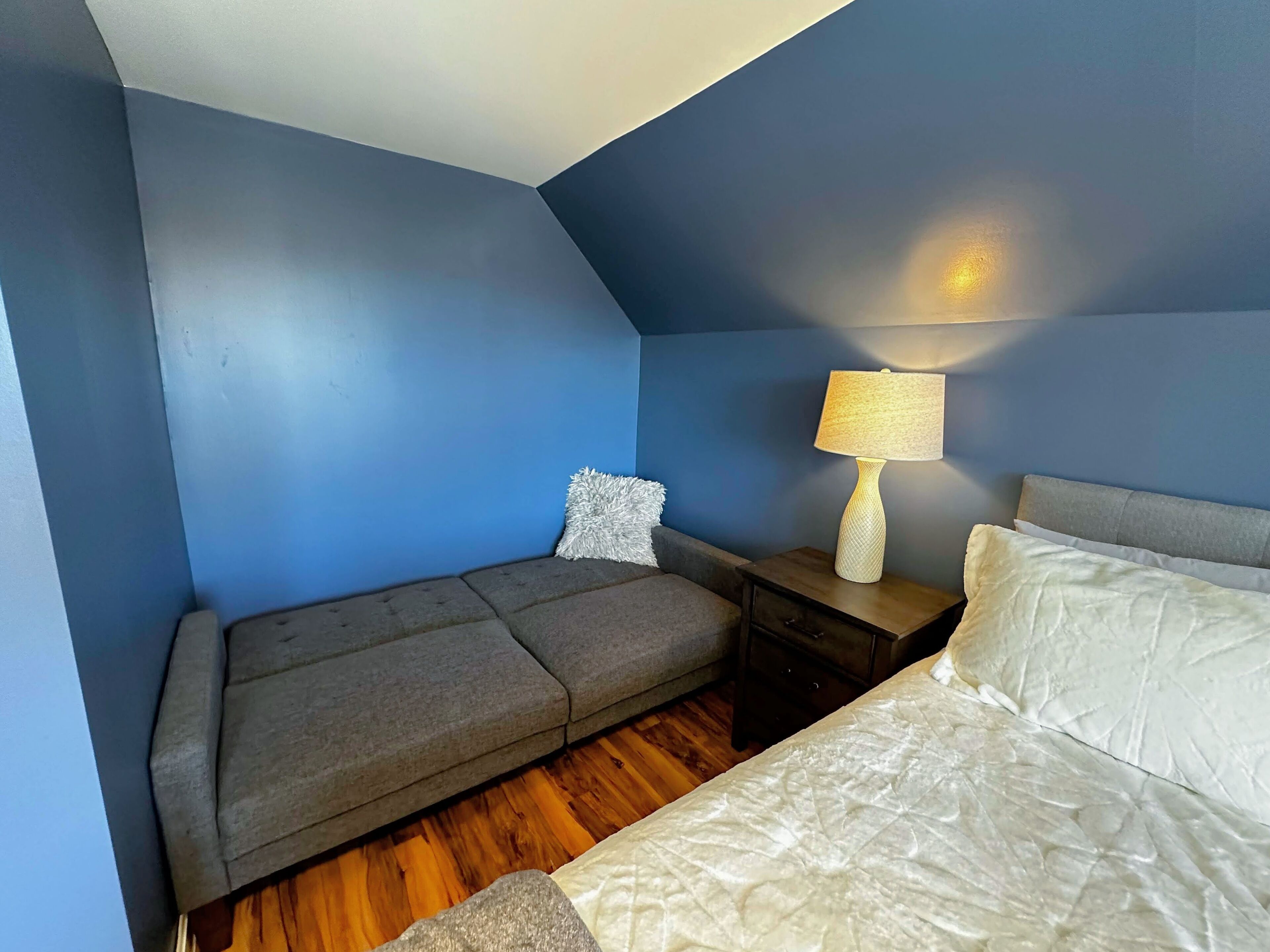 Une pièce avec un canapé gris, un lit blanc, une table de chevet avec une lampe et un mur bleu.