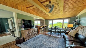 A spacious living room with a wooden ceiling, a large TV, a leather sofa, and a dining area with a view of the outdoors. - 64J Stunning sunsets, just 20 mins to Bretton Woods. Hot Tub, Foosball, AC, dog friendly! (Whitefield)