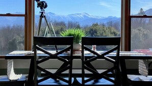 A room with a telescope on a tripod, a table with chairs, and a view of snow-capped mountains through large windows. - 64J Stunning sunsets, just 20 mins to Bretton Woods. Hot Tub, Foosball, AC, dog friendly! (Whitefield)