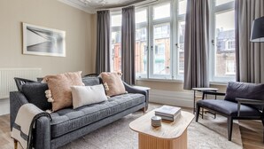 Una sala de estar moderna con un sofá gris, una mesa de centro de madera y un sillón negro. Hay ventanas grandes con cortinas, un radiador y un cuadro en la pared.
