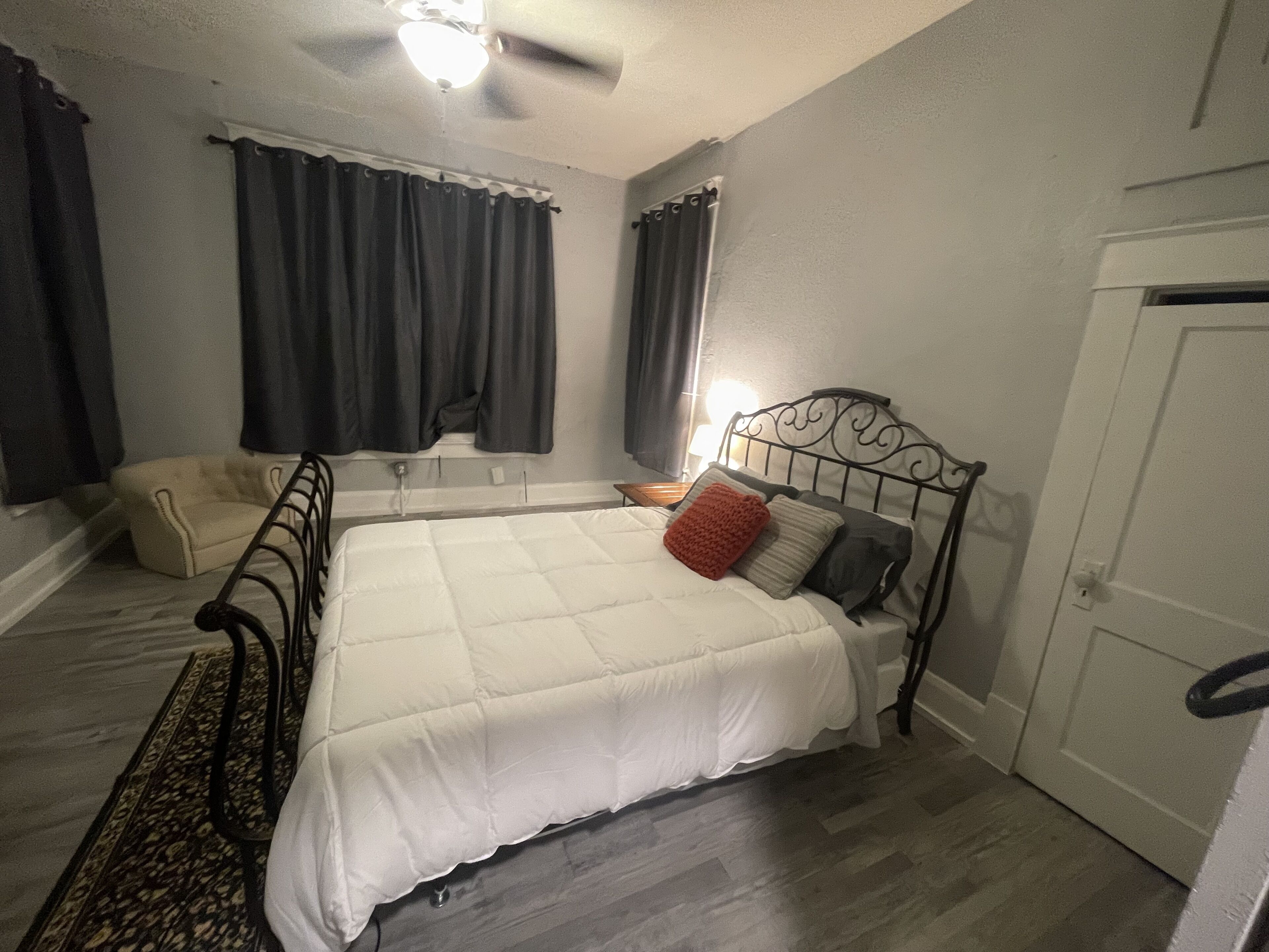 Une chambre à coucher comprenant un lit, une chaise, une fenêtre avec des rideaux, un ventilateur de plafond et une porte.