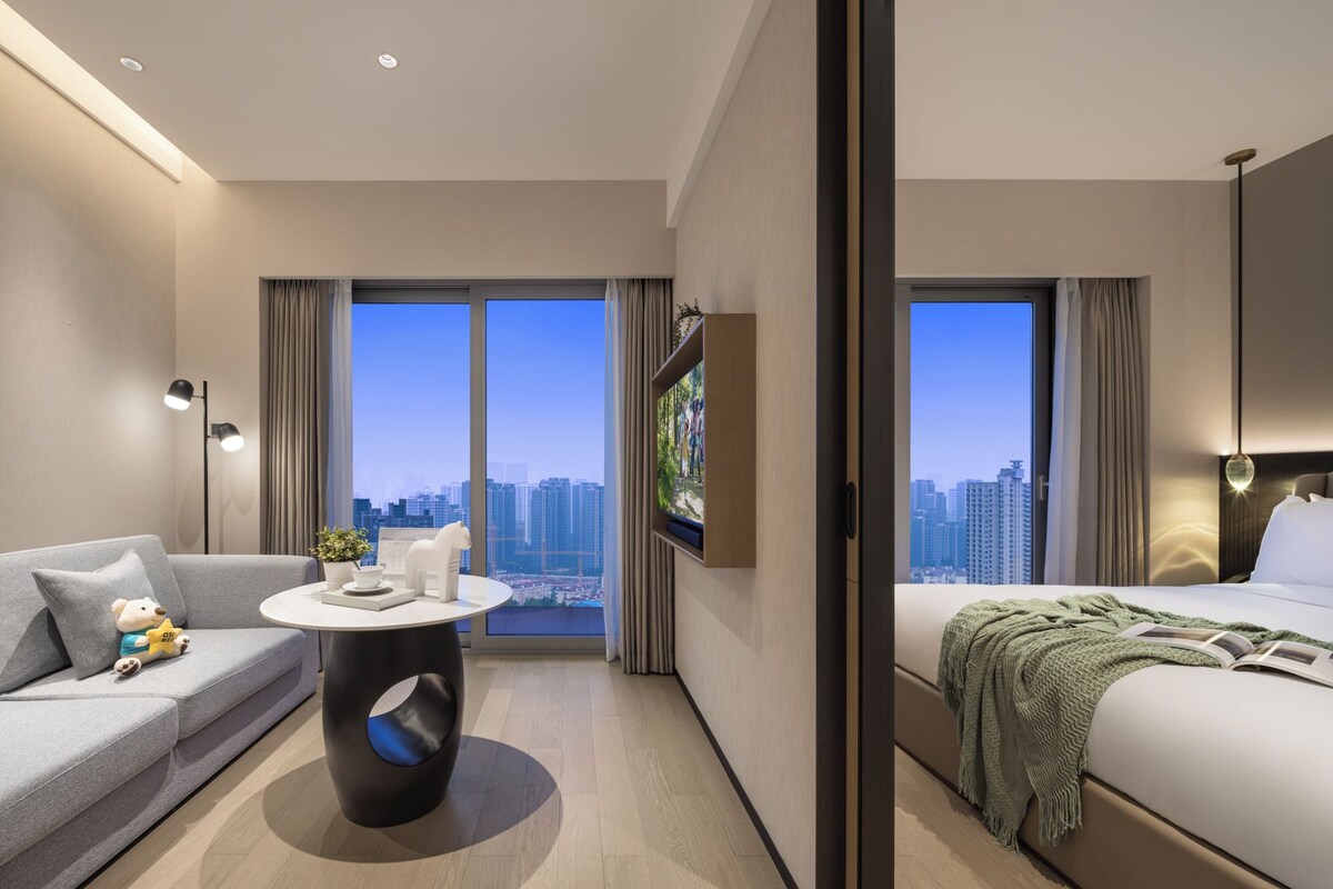 A modern hotel room with a sofa, a round table, a bed, and a large window offering a city view.