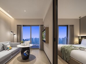 A modern hotel room with a sofa, a round table, a bed, and a large window offering a city view.