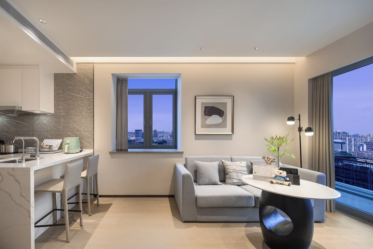 A modern apartment with a kitchen, a dining area, a living room with a sofa and a coffee table, a large window with a city view, and a framed artwork on the wall.