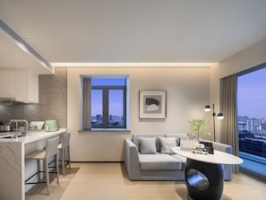 A modern apartment with a kitchen, a dining area, a living room with a sofa and a coffee table, a large window with a city view, and a framed artwork on the wall.