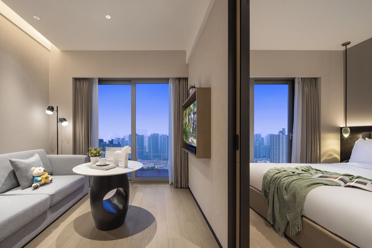 A modern hotel room with a sofa, a round table, a bed, and a large window offering a city view.