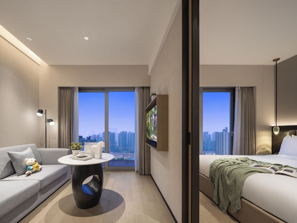 A modern hotel room with a sofa, a round table, a bed, and a large window offering a city view.