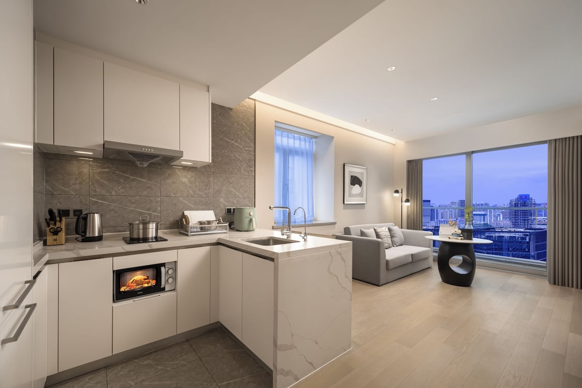 A modern kitchen with a built-in oven, a dining area with a sofa, and a large window offering a city view.