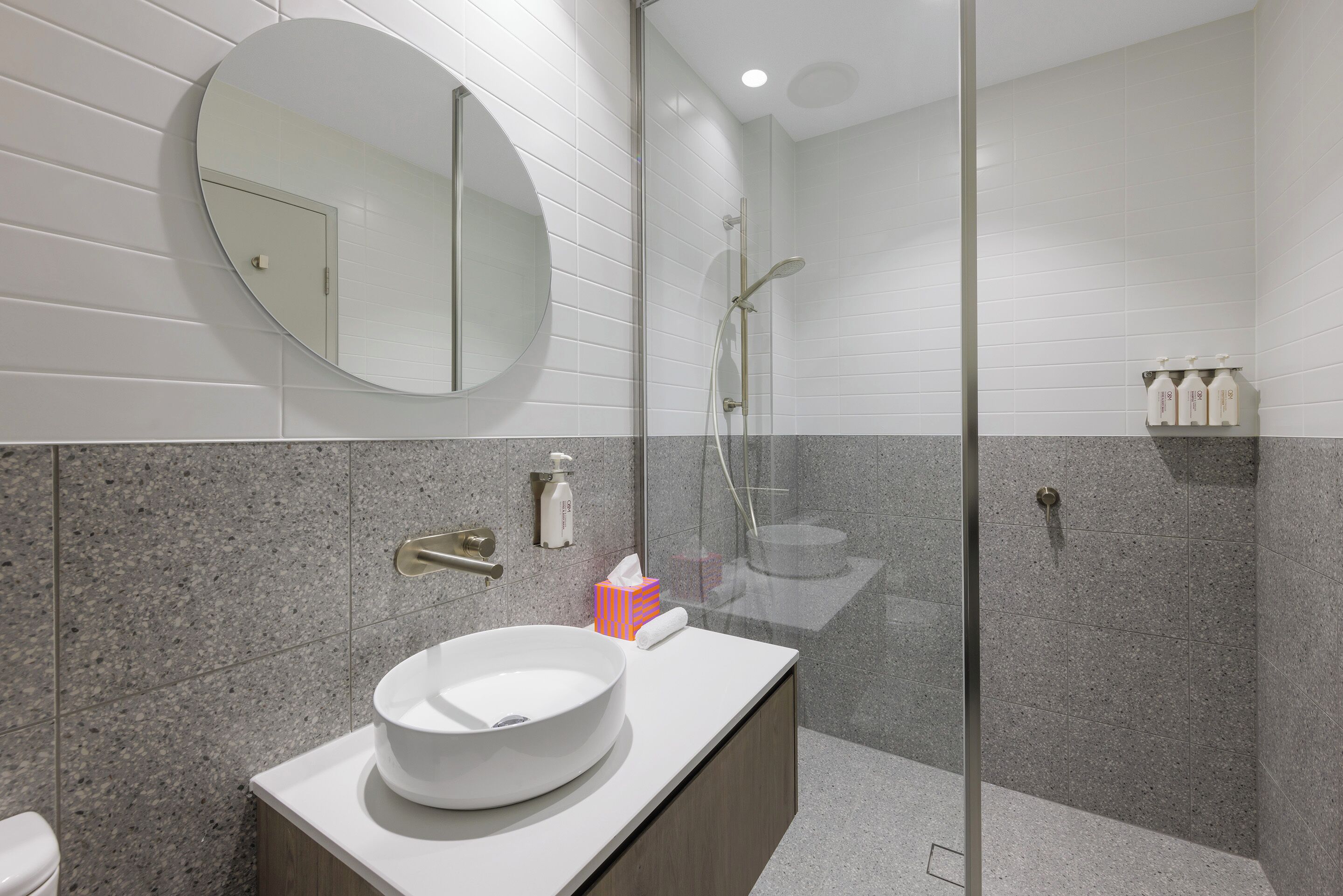 Un baño moderno con un espejo redondo, un lavabo blanco y una zona de ducha con mampara de vidrio.