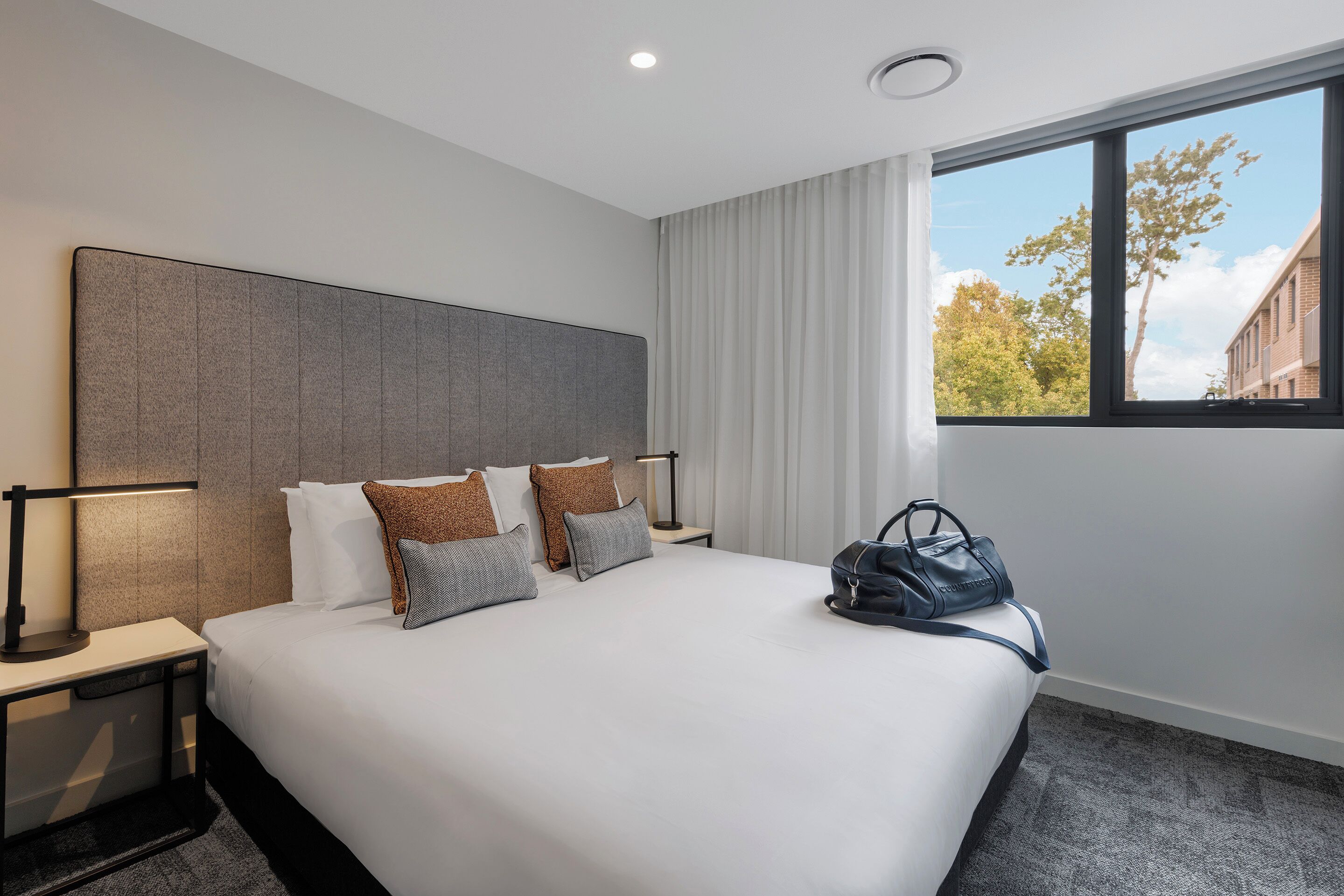 A neatly made hotel room with a large bed, a bedside table with a lamp, and a view of trees through a window.