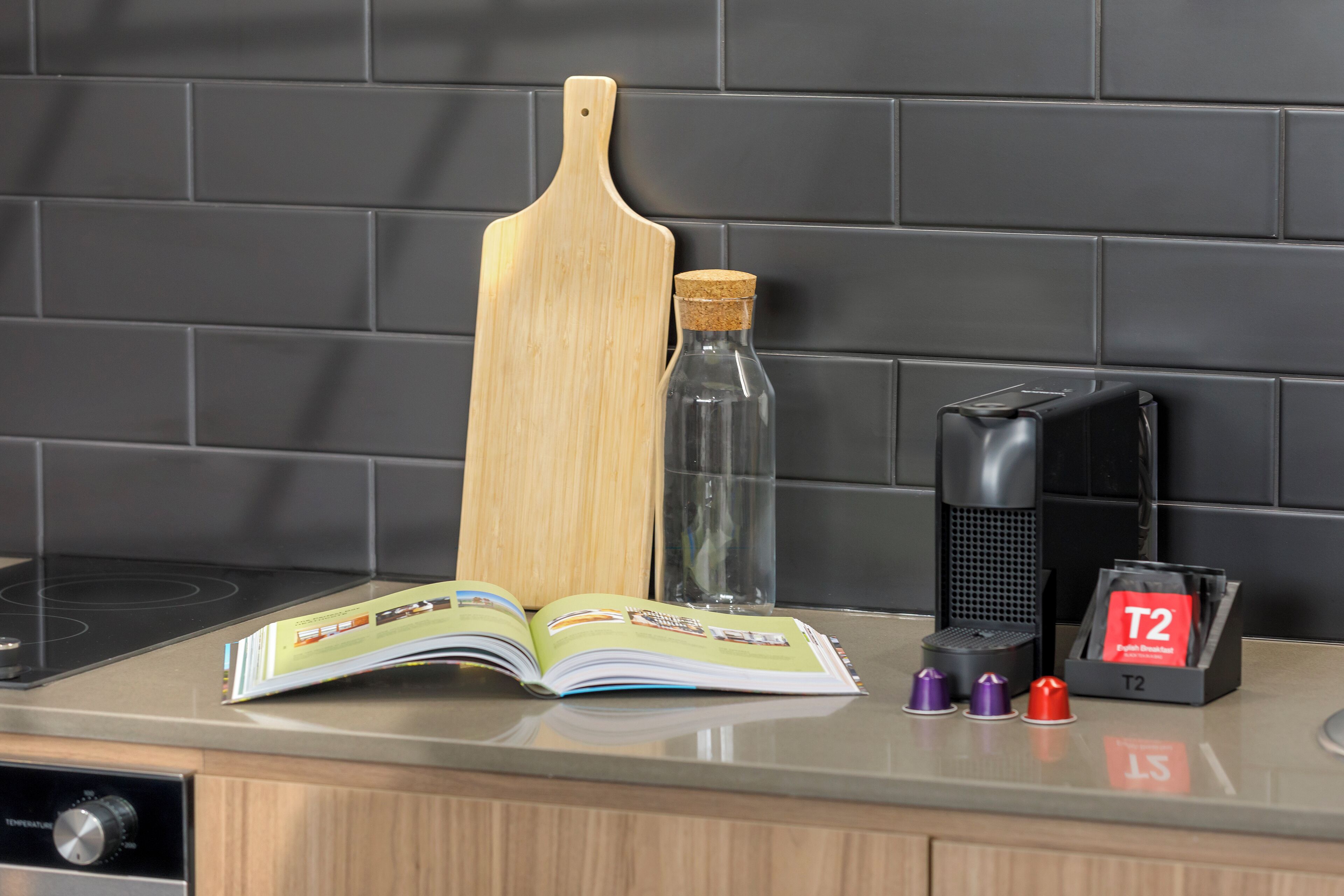 A kitchen counter with a wooden cutting board, a glass bottle, a coffee maker, and a T2 branded coffee pod holder.