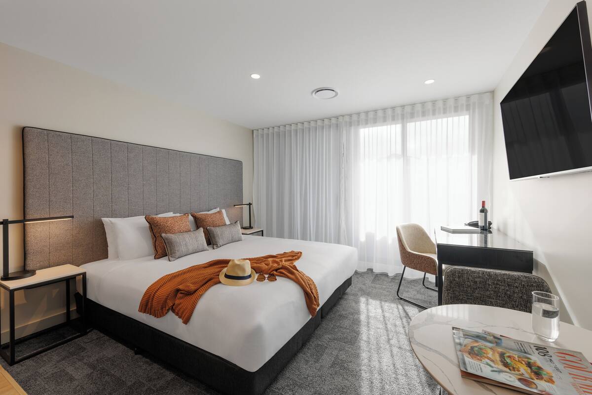 A modern hotel room with a large bed, a desk, a chair, a TV, and a magazine on the table.