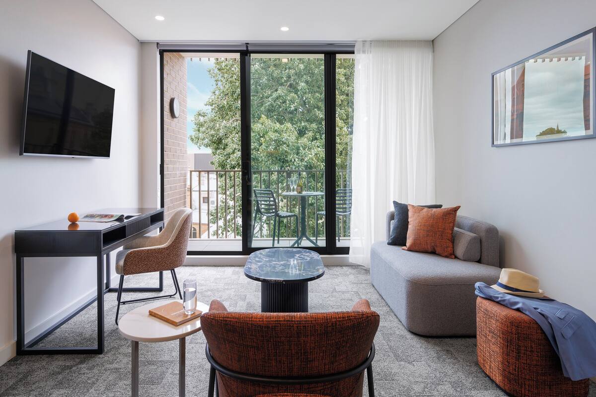 A modern room with a desk, chair, sofa, and a balcony with outdoor seating.