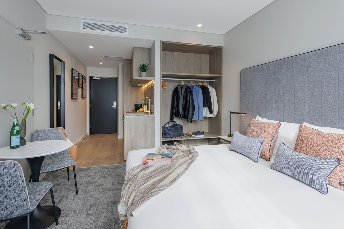 A modern hotel room with a large bed, a walk-in closet, a dining area with a table and chairs, and a small kitchen area.
