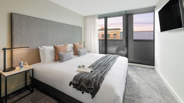 A modern hotel room with a large bed, a flat-screen TV, a bedside table with a lamp, and a view of the city through the window. - Punthill Maitland (Maitland)