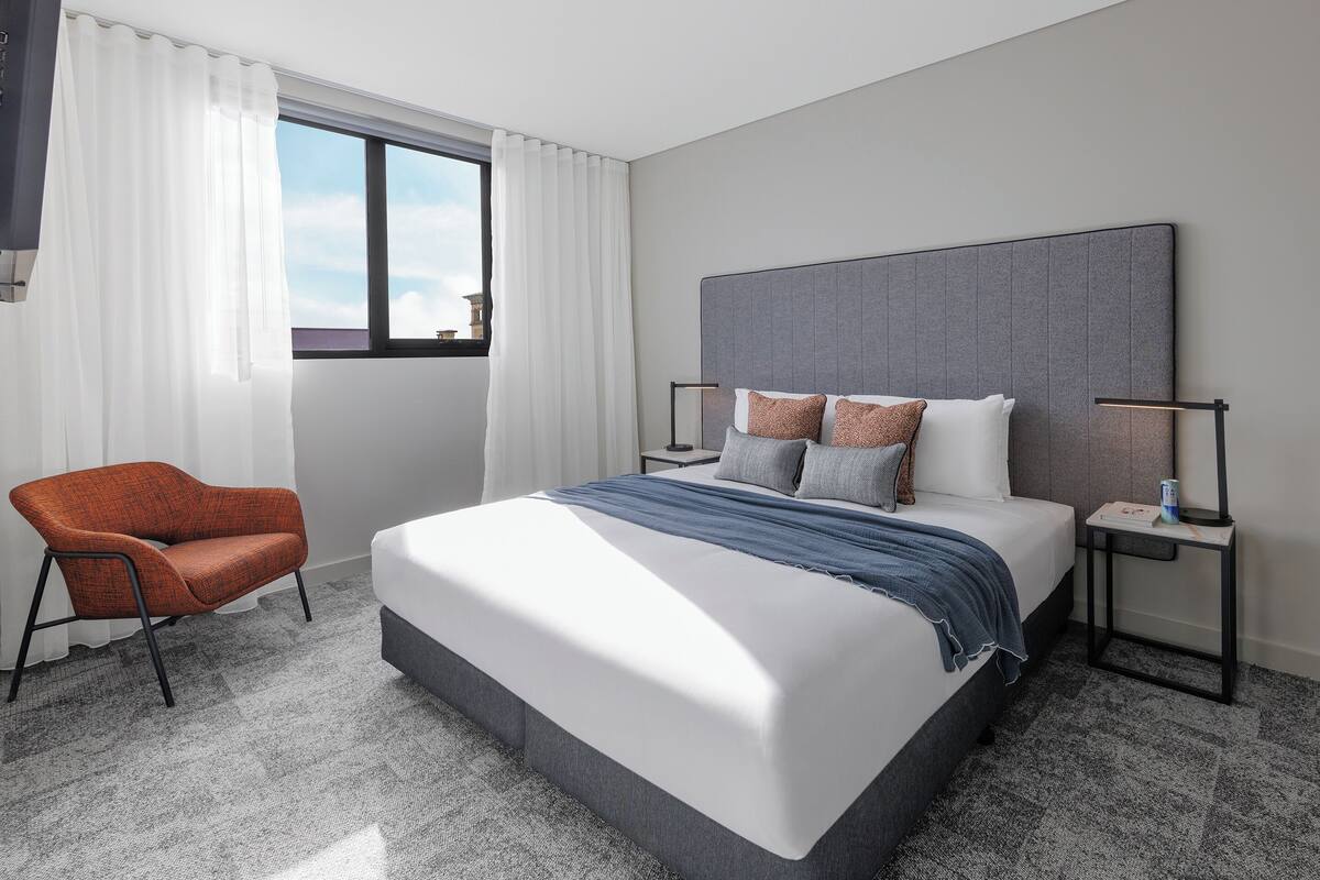 A modern hotel room with a large bed, a chair, a nightstand, and a window with curtains.