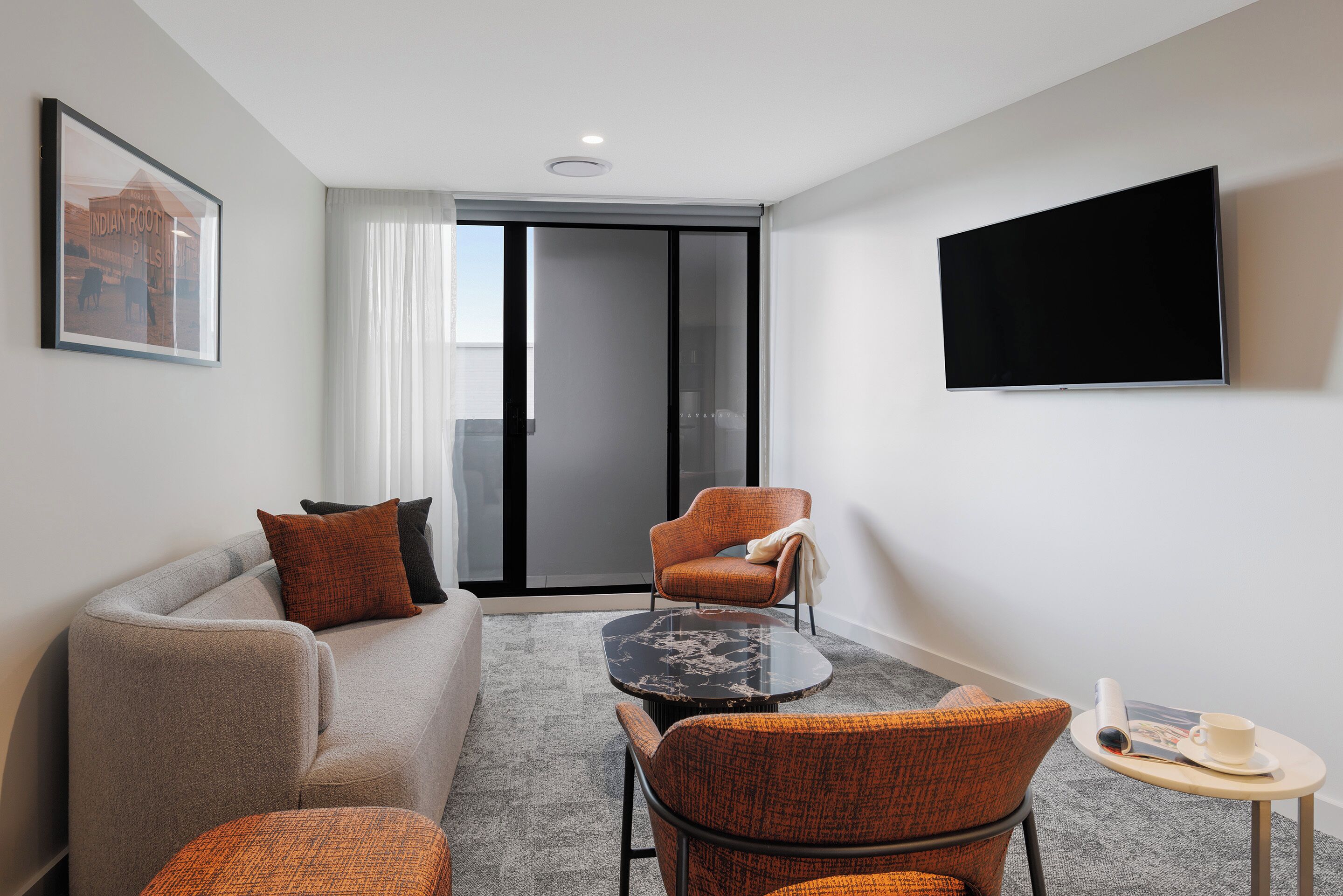 Ein modernes Hotelzimmer mit grauem Sofa, orangenen Sesseln, einem kleinen runden Tisch und einem Flachbildfernseher an der Wand.