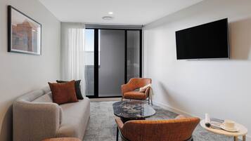 Ein modernes Hotelzimmer mit grauem Sofa, orangenen Sesseln, einem kleinen runden Tisch und einem Flachbildfernseher an der Wand.