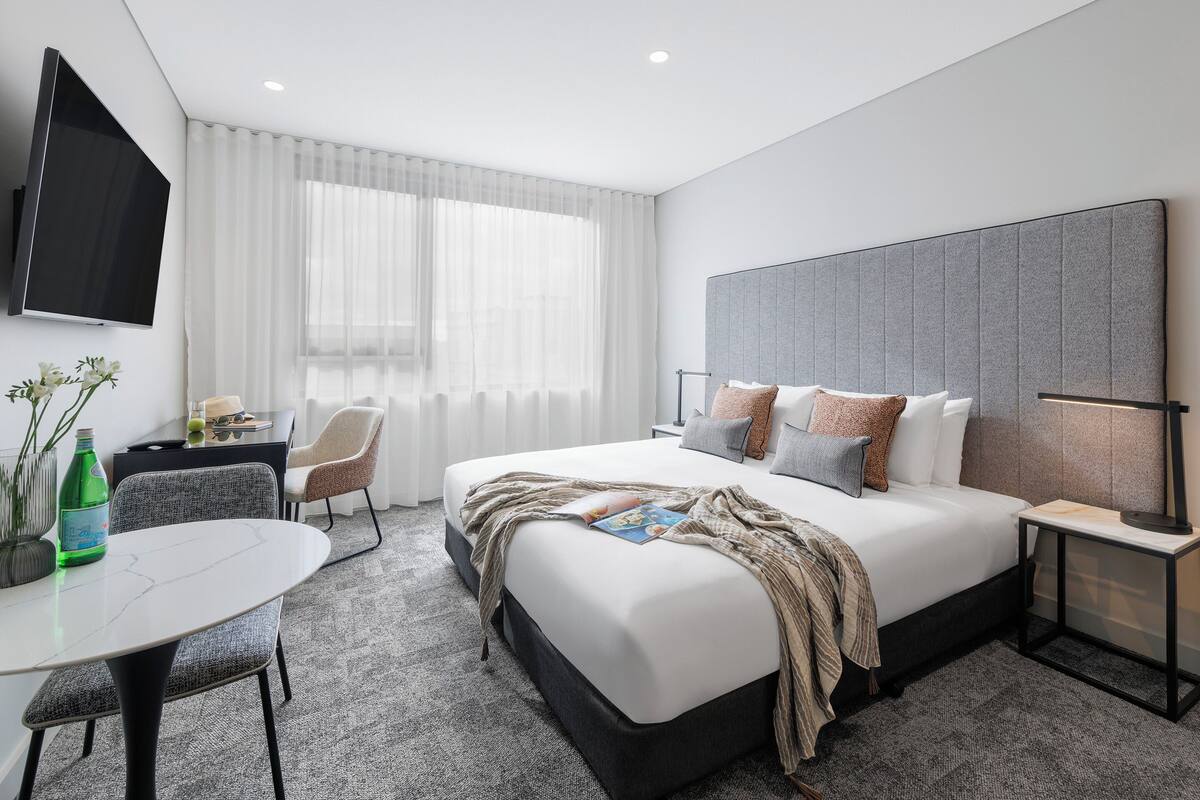 A modern hotel room with a large bed, a desk, a chair, a TV mounted on the wall, and a window with sheer curtains.