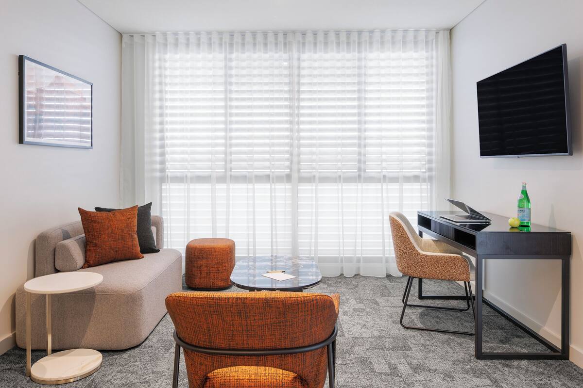 A modern hotel room with a sofa, two armchairs, a small round table, a desk with a computer, and a large window with blinds.