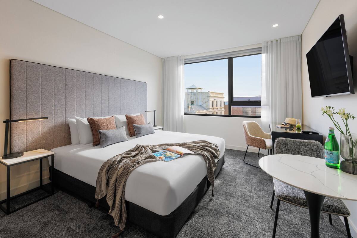 A modern hotel room with a large bed, a desk, a chair, a TV, and a window with a city view.