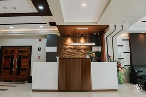 Una recepción con diseño moderno, que incluye un panel de madera y una barra blanca. Hay luces en el techo sobre la barra, y se ve una planta a la derecha.