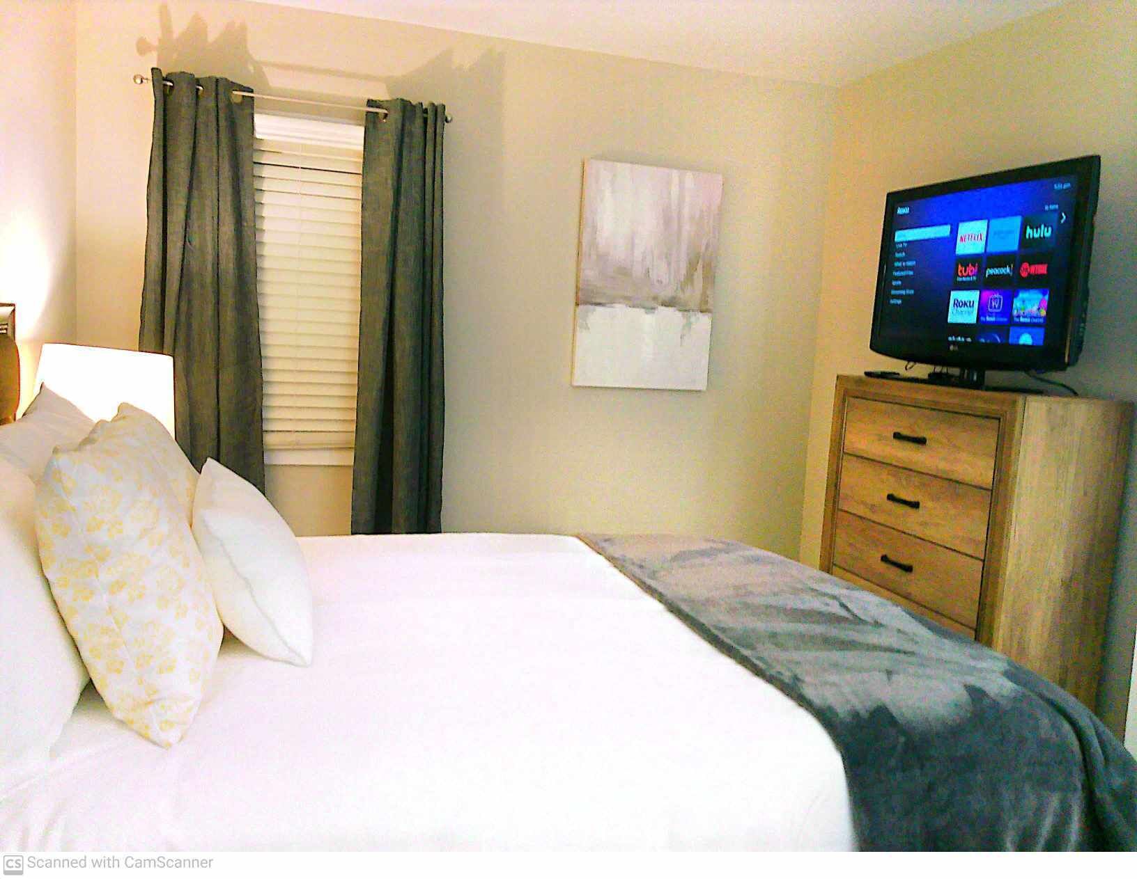 A hotel room with a bed, a television displaying streaming services, a painting on the wall, and a wooden dresser.