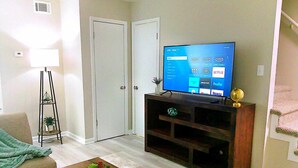 A living room with a flat-screen TV on a wooden stand, a sofa, a coffee table with a plant, a floor lamp, and a door.