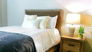 A neatly made bed with white linens and a dark throw blanket, a bedside lamp, a small potted plant, and a wooden nightstand with drawers.