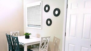 A dining area with a white table and chairs, a potted plant, and a window with blinds.