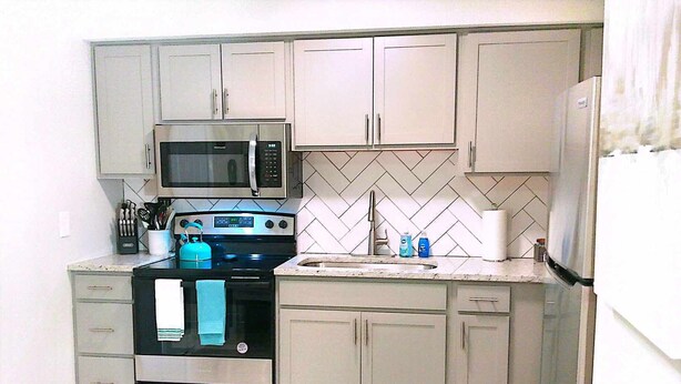 A compact kitchen with a microwave, stove, and sink. It includes cabinets, a refrigerator, and a tiled backsplash.