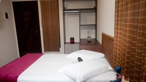Quarto de hotel com uma cama, penteadeira, espelho e uma pequena mesa com um objeto decorativo.