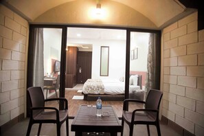 A hotel room with a bed, a desk, a chair, a table, and a sliding glass door leading to a balcony.