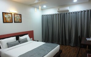 A hotel room with a bed, a nightstand, a desk, a chair, a window with curtains, an air conditioning unit, and two wall-mounted paintings. - Hotel Pearls (Chhatrapati Sambhajinagar)