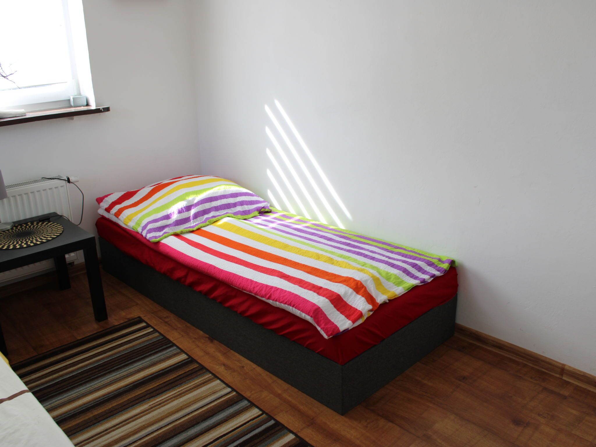 Ein kleines Zimmer mit einem Einzelbett, einer gestreiften Bettdecke, einem roten Kopfkissen und einem schwarzen Nachttisch mit einem Dekorationsgegenstand.
