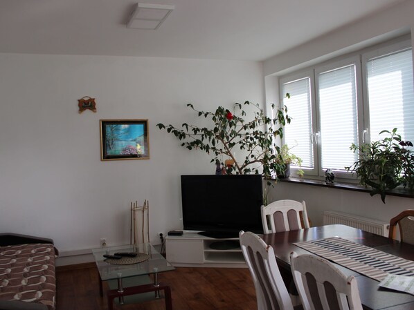 A living room with a TV, a glass coffee table, a dining table with chairs, a potted plant, and a painting on the wall.