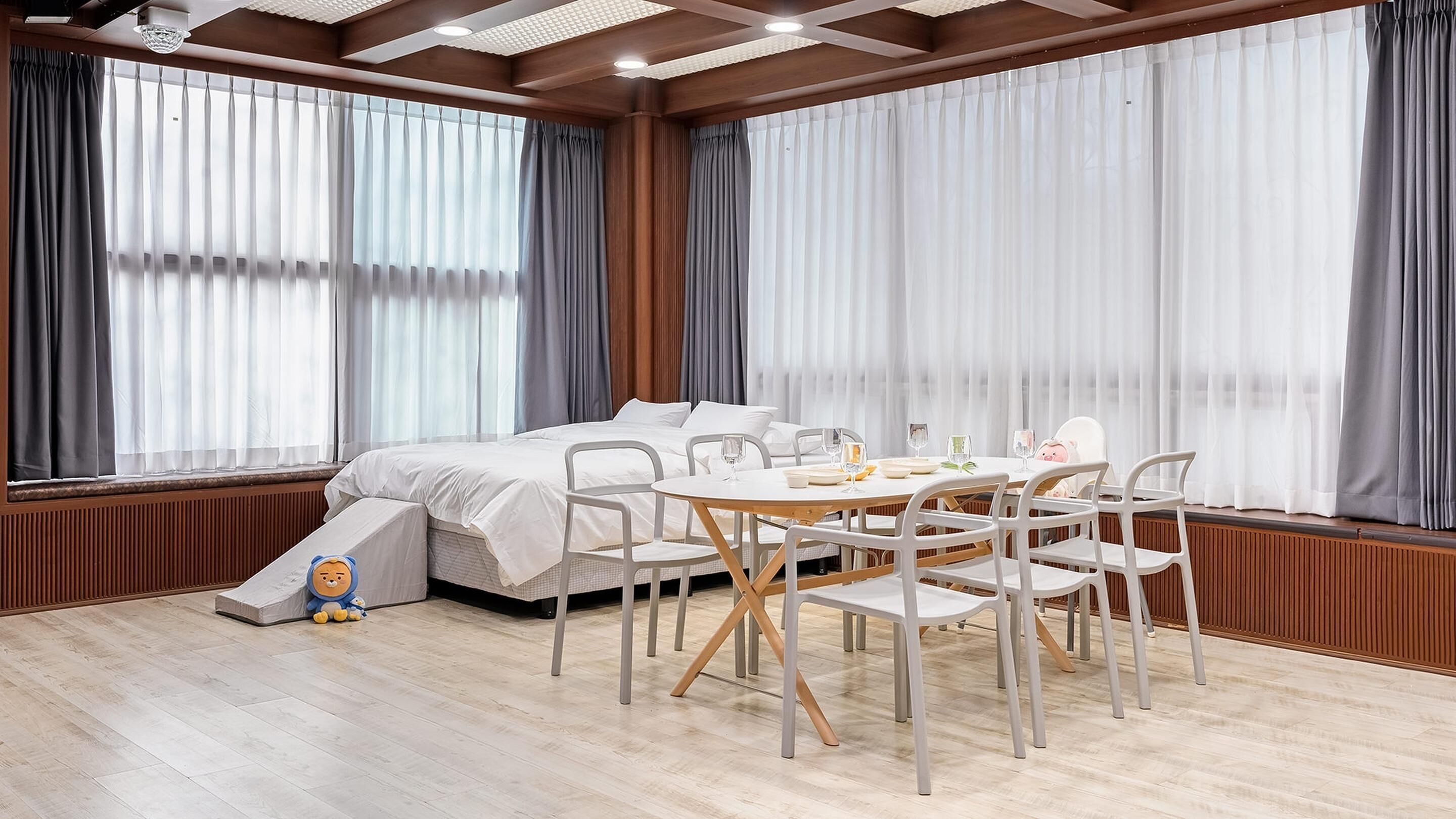 Una habitación de hotel con una cama, una mesa de comedor con sillas y ventanas amplias con cortinas.
