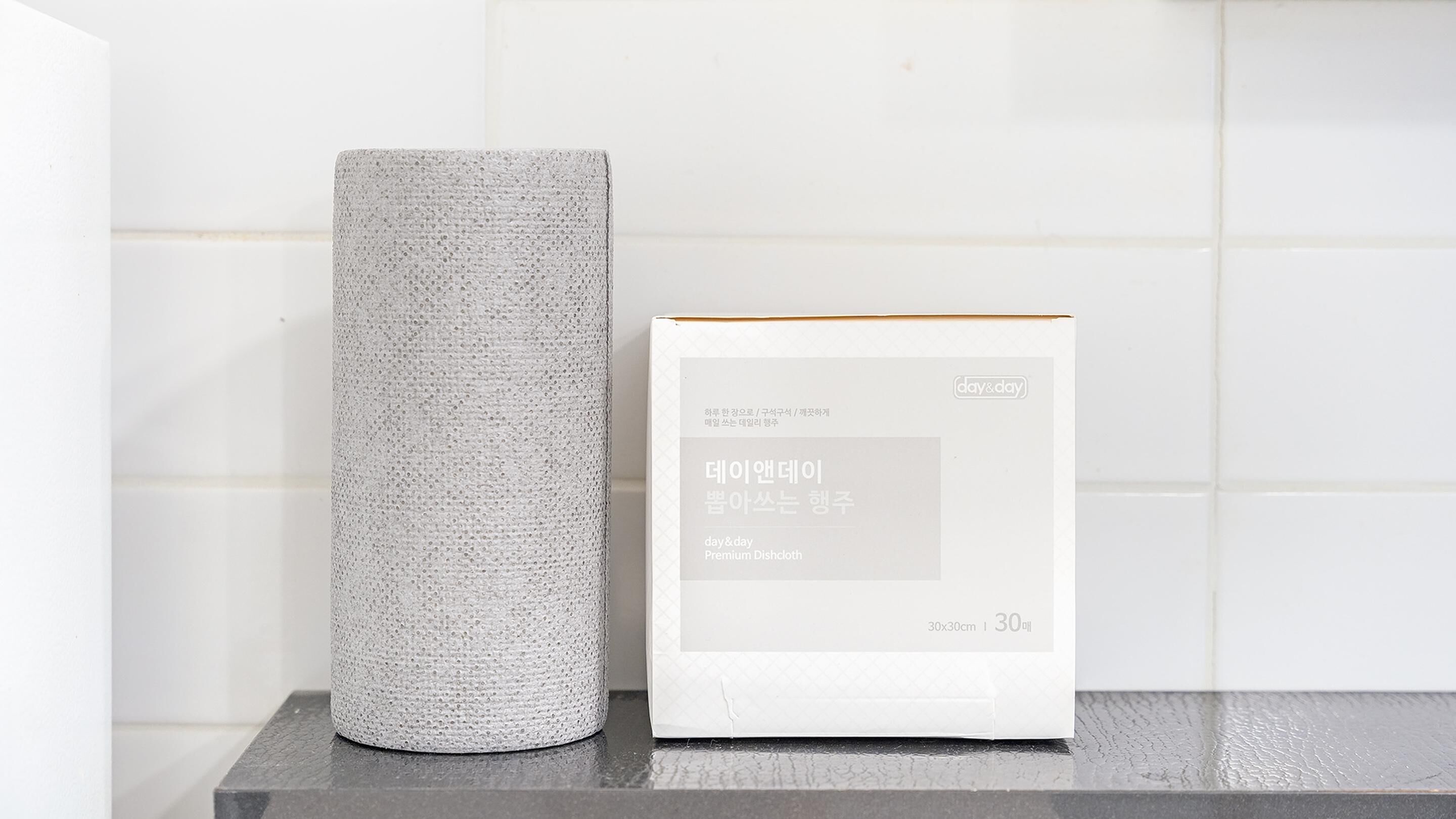 A white dishcloth and a box of day&day Premium Dishcloth on a dark surface against a white tiled wall.