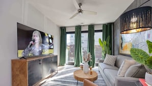 Una sala de estar moderna con televisor de pantalla plana, ventilador de techo, sofá, mesa de centro y un ventanal con cortinas.