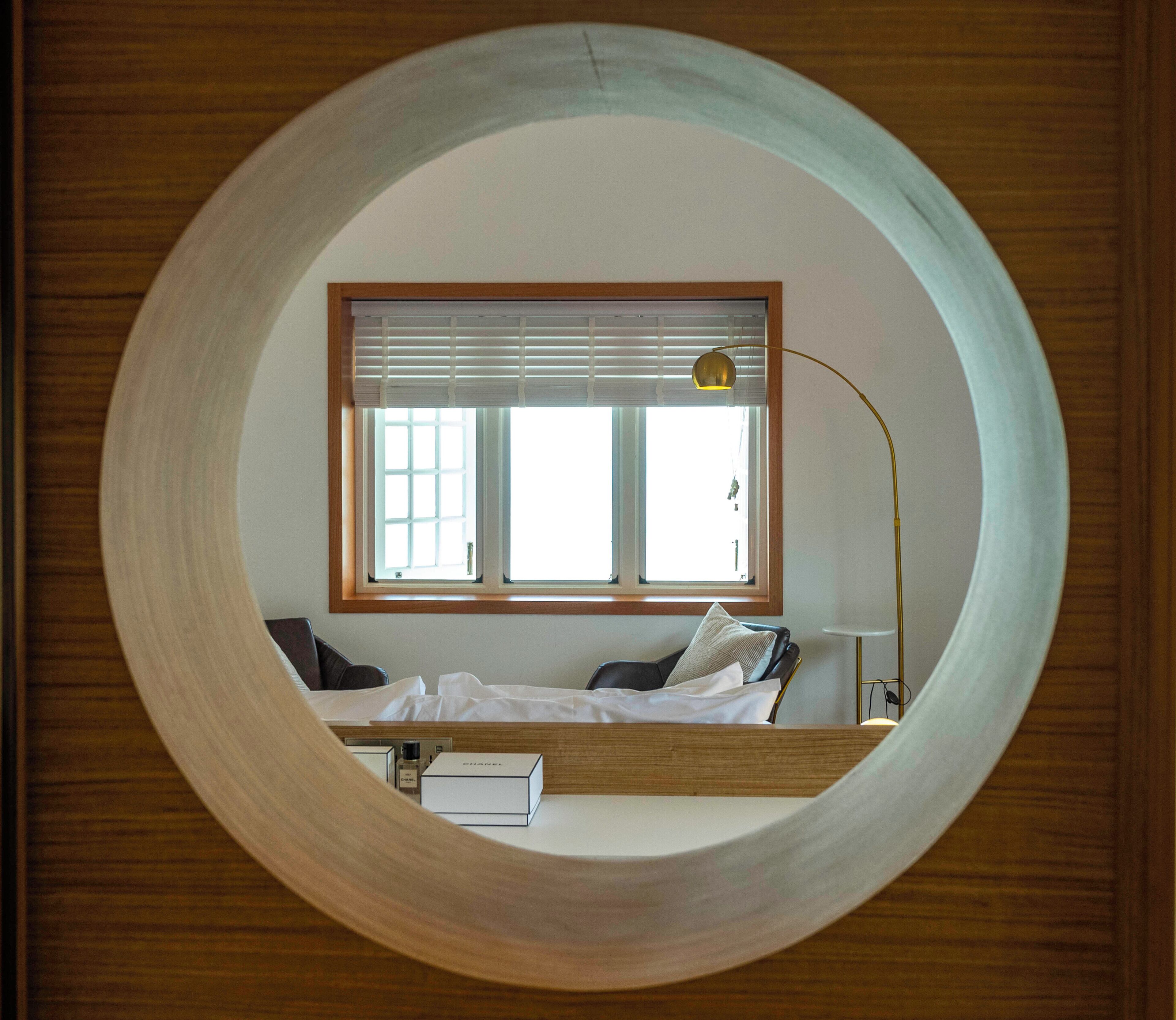 A circular window framing a bed with a window above, a lamp, and a bedside table.