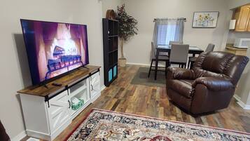 Una sala de estar con televisor, sillón de cuero, mesa de comedor y cocina al fondo.