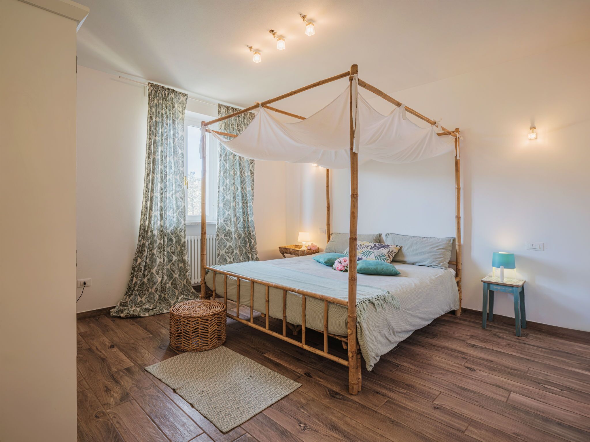 A bedroom with a four-poster bed, a small bedside table, a nightstand, a wicker basket, and a floor rug.