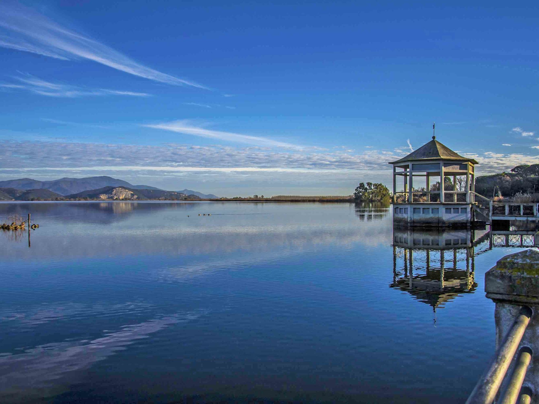 Ein friedlicher See mit einem Pavillon auf Stelzen, umgeben von ruhigem Wasser und fernen Bergen.